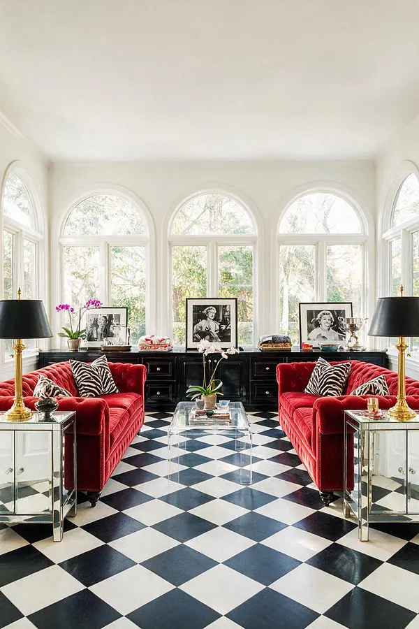 A bright sunroom with tall arched windows, black-and-white checkerboard flooring, and two deep red velvet sofas facing a clear acrylic coffee table. Mirrored side tables, black lamps with gold bases, and a long black cabinet with framed black-and-white portraits create a glamorous Hollywood Regency look.