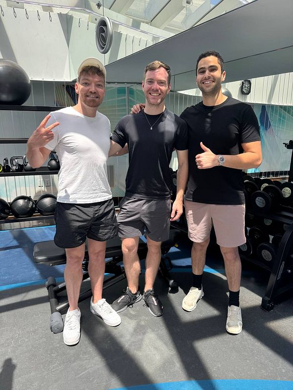Three men pose together in a gym setting, smiling and showing friendly gestures.