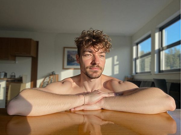 A man with curly hair is sitting at a table in a sunlit room.