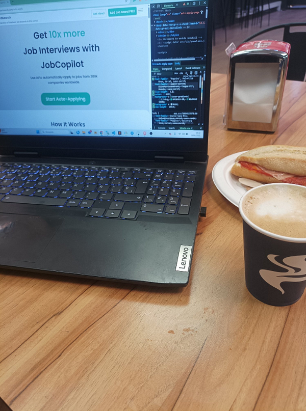 A laptop is set up on a wooden table with a coffee cup and a sandwich nearby.
