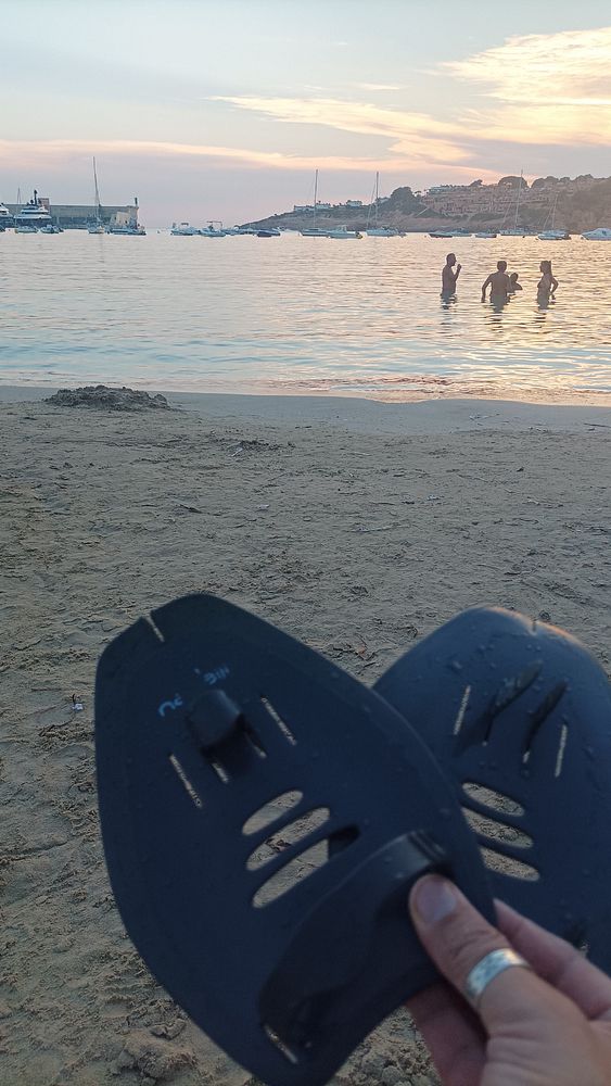 A person holds swim fins in front of a serene beach scene at sunset.