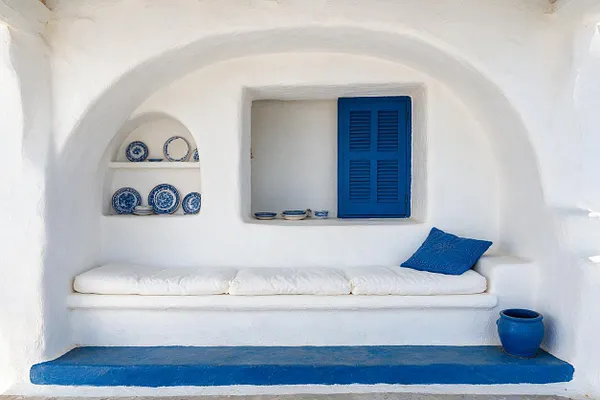 A serene Mediterranean-style front porch with smooth white stucco walls, a rounded arched ceiling, and built-in niches displaying blue-and-white ceramic plates. A white cushioned bench spans the wall beneath a bright blue shutter, with a blue throw pillow and a cobalt floor step and planter adding bold contrast.