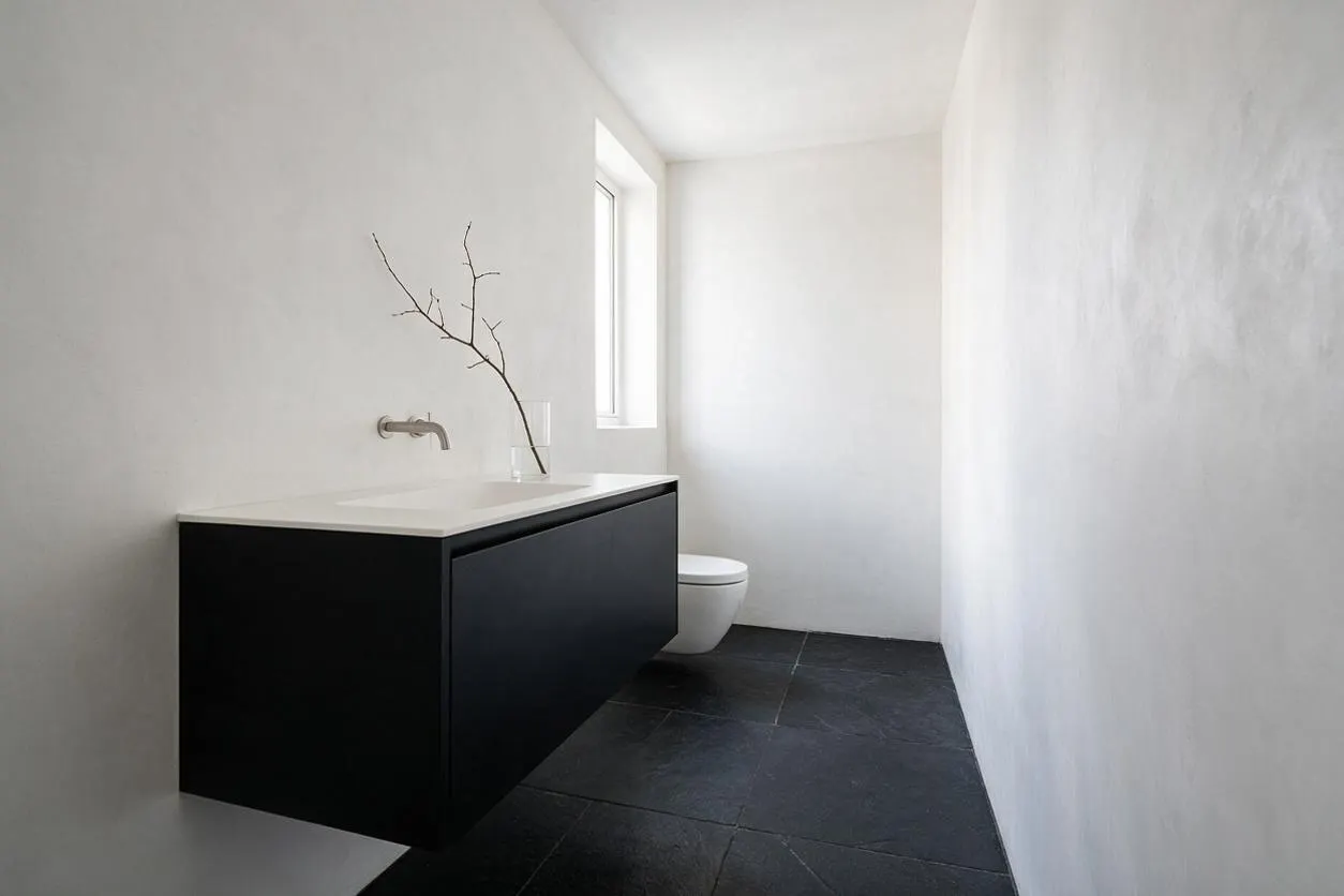 A narrow minimalist powder room with smooth white walls, a floating matte black vanity, and a white countertop. Dark stone floor tiles contrast with the bright space, while a small window and a simple branch in a vase add a sparse decorative touch.