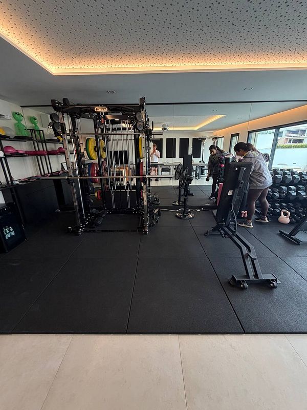 A photograph of a gym setup with exercise machines, dumbbells, and a roller barrel on a wooden floor against a blue painted wall.