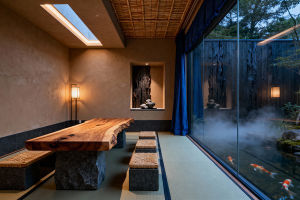 A Japanese Zen dining room features a low live-edge wood table on rough stone bases with woven stools atop tatami mats. Earthen plaster walls and a bamboo ceiling frame a niche with charred timber and river stones, lit by a slender floor lantern and a linear skylight. A full-height glass wall with indigo drapes overlooks a misted koi pond and dark wood garden wall, creating a calm, minimalist atmosphere.