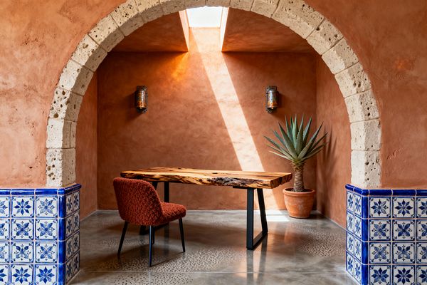 A Southwestern-inspired home office sits within a terracotta plaster alcove framed by a pale stone arch and blue-and-white Talavera tile. A live-edge wood desk on black steel legs pairs with a rust boucle chair, while an agave in a terra-cotta pot and two hammered copper sconces flank the back wall; a skylight above sends a bright beam across the plaster and polished concrete floor, creating a warm, focused workspace.