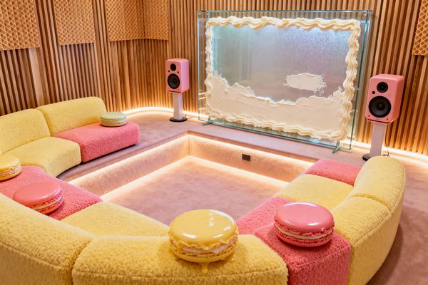 A sunken media room centers on a curved modular sofa upholstered in fluffy yellow and pink boucle, framing a carpeted pit with soft perimeter lighting. Glossy macaron-shaped stools rest on the cushions, pink speakers on white stands flank a glass screen trimmed in frosting-like edging, and vertical oak slat walls with waffle-pattern acoustic panels glow warmly, reinforcing the playful Candy Land palette and cozy, acoustically treated atmosphere.