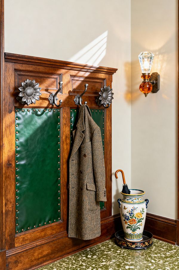 A Victorian-style mudroom corner with a stained oak hall tree featuring emerald green leather panels edged in brass nailhead trim. Ornate pewter sunflower rosettes and curved metal hooks hold a tweed overcoat. To the right, a wall sconce with a faceted crystal shade and amber glass globe casts warm light on cream walls. A hand-painted ceramic umbrella stand on a mottled green terrazzo floor contains a black umbrella with a wooden handle.