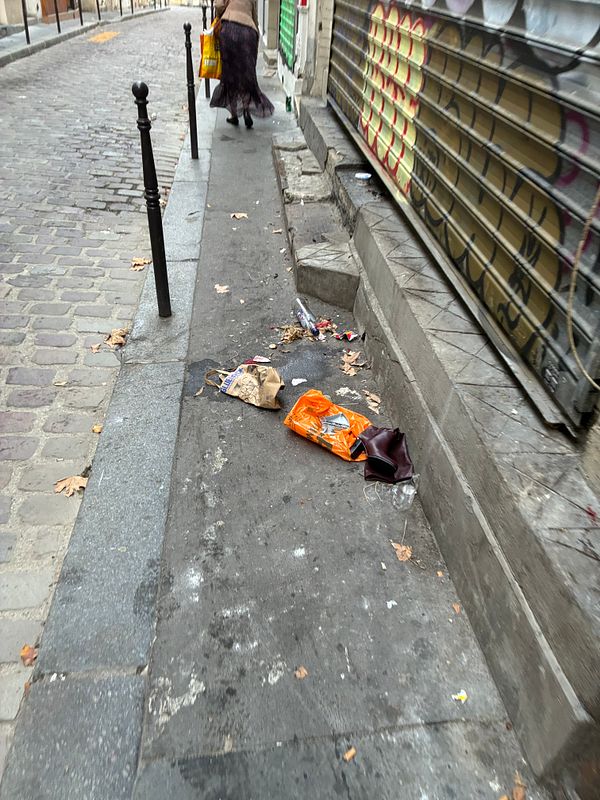 Narrow urban sidewalk in Paris littered with trash next to a graffiti-covered metal shutter and a person walking away carrying a yellow bag.