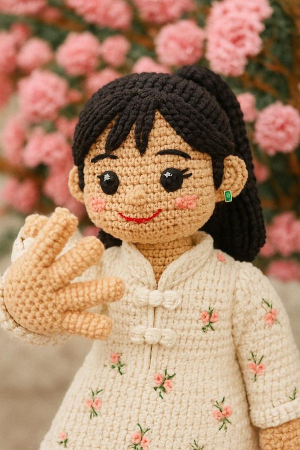 A crocheted doll with a friendly expression stands in front of a backdrop of pink flowers.