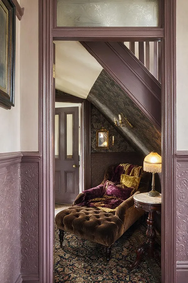 A narrow Victorian basement room with a sloped ceiling, dark patterned wallpaper, and rich plum trim. A brown velvet chaise lounge with jewel-toned pillows sits beside a small round marble-topped table and fringed lamp, while a floral rug anchors the space.