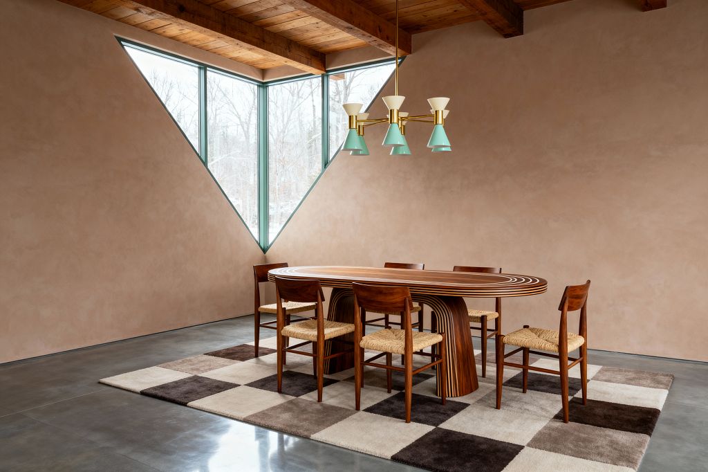 A minimalist dining room centers on a striped oval wood table with waterfall legs, surrounded by six walnut chairs with woven rush seats on a cream and dark-brown checkered rug. Warm plaster walls, a triangular corner window with sage-green frames, exposed timber ceiling, polished concrete floor, and a brass chandelier with mint and white cone shades complete the mid-century modern composition.