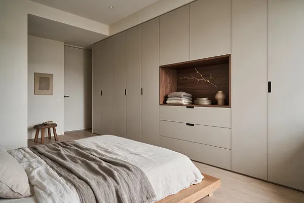 A serene Japandi-style closet room with floor-to-ceiling beige built-in wardrobes, a wood-lined display niche, and a low platform bed in the foreground. Soft white bedding, a textured throw, and minimal decor create a calm, warm atmosphere.