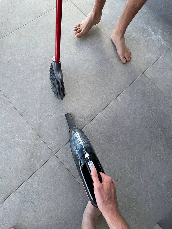 The image captures a person cleaning a tiled floor using a vacuum cleaner and a broom.