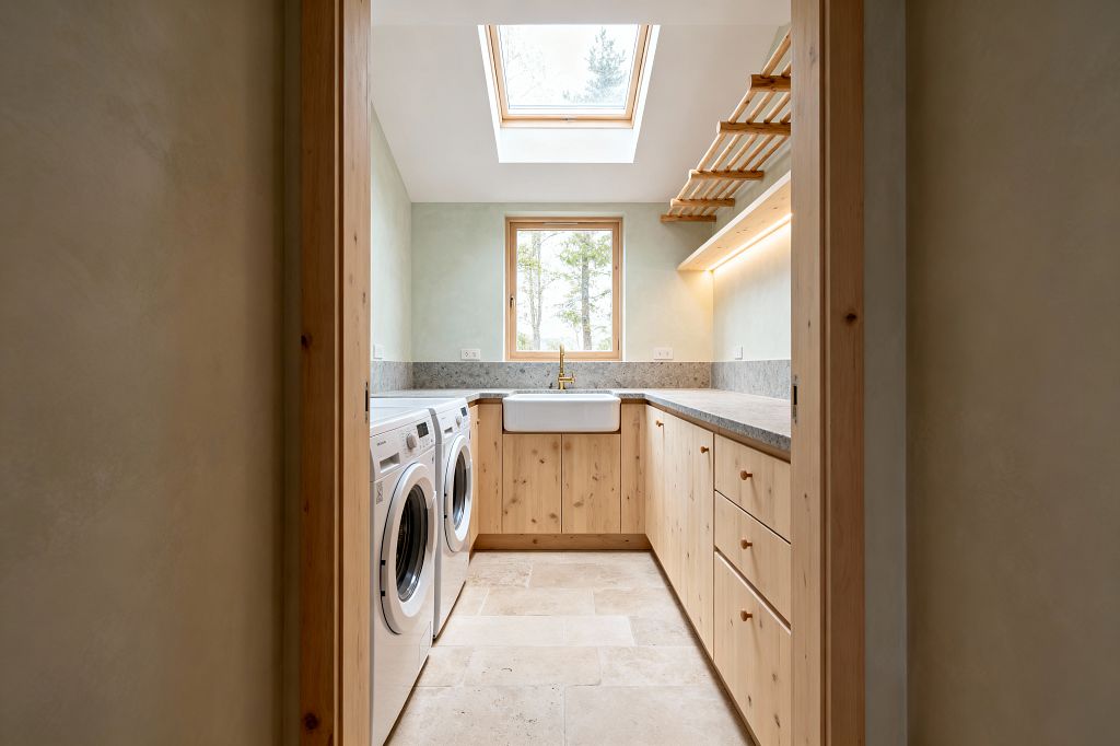 A narrow Scandinavian-style laundry room with knotty pine base cabinets and round wood knobs forming a compact U-shaped layout. A white apron-front sink with a brass faucet sits under a wood-framed window, while a skylight floods the space with daylight. Gray stone-look countertops, a slim backsplash, and white front-loading washer and dryer line the left side. Pale green limewashed walls, a ceiling-mounted wooden drying rack, warm under-cabinet lighting, and beige limestone floor tiles complete the scene.