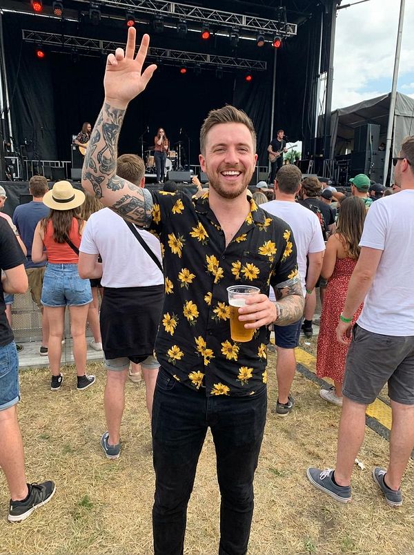 A man at a music festival is smiling and raising his hand while holding a drink.