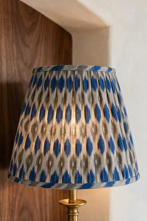A close-up of a table lamp with a pleated fabric lampshade in blue, beige, and gray ikat-like pattern, mounted on a brass stem. The lamp sits against a backdrop of warm wood paneling and a white wall, creating a layered eclectic look.