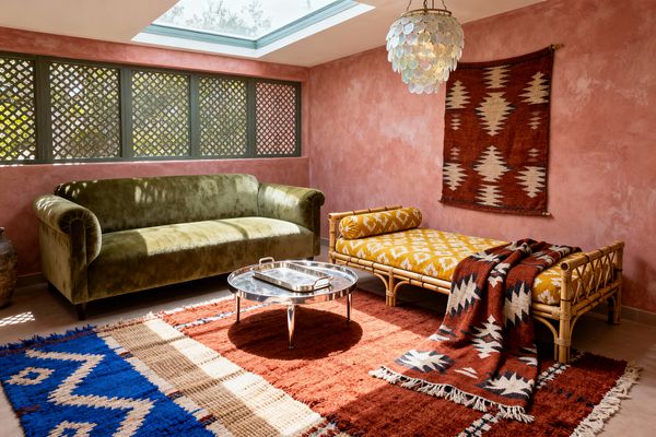 A Bohemian living room features an olive green velvet sofa opposite a rattan daybed with mustard ikat cushions, set on layered wool kilim rugs in rust, cream, and cobalt. Textured pink plaster walls surround green lattice windows and a skylight, with a capiz shell pendant overhead, a rust geometric wall hanging, a low polished metal round coffee table with a tray at center, and a large earthenware pot in the corner, all bathed in warm natural light.