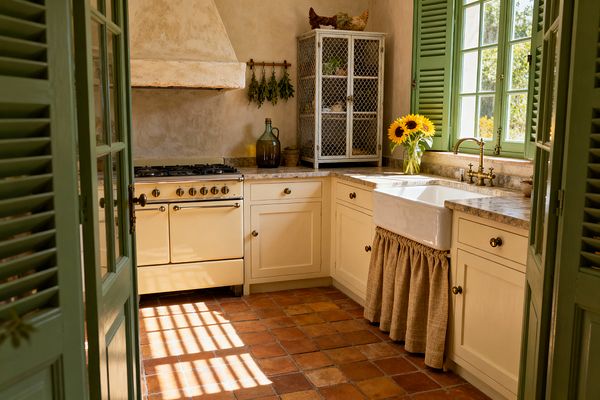 Sunlit French country kitchen with sage-green shutters and doors, cream cabinets, and a vintage-style cream range beneath a plaster hood. Terracotta floor tiles lead to a porcelain farmhouse sink with a gathered burlap skirt and a brass faucet set into beige stone countertops. A wire-mesh hutch, dried herbs, a green glass demijohn, and a vase of sunflowers add rustic character and a lived-in feel.