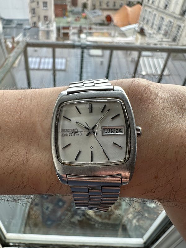 A vintage Seiko automatic watch from the 1970s is displayed on a wrist with a cityscape in the background.