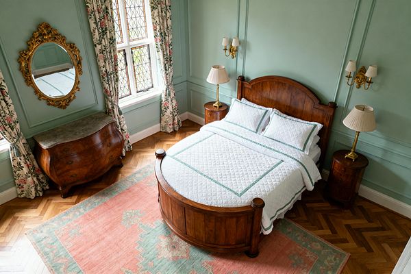 Overhead view of a traditional bedroom with mint-green paneled walls and a walnut bed featuring an arched headboard and curved footboard. White quilted bedding with green trim sits atop a coral and sage area rug on herringbone parquet, flanked by round wooden nightstands with brass lamps, brass wall sconces, floral chintz curtains at a leaded window, and a gilt oval mirror above a marble-topped bombe chest.