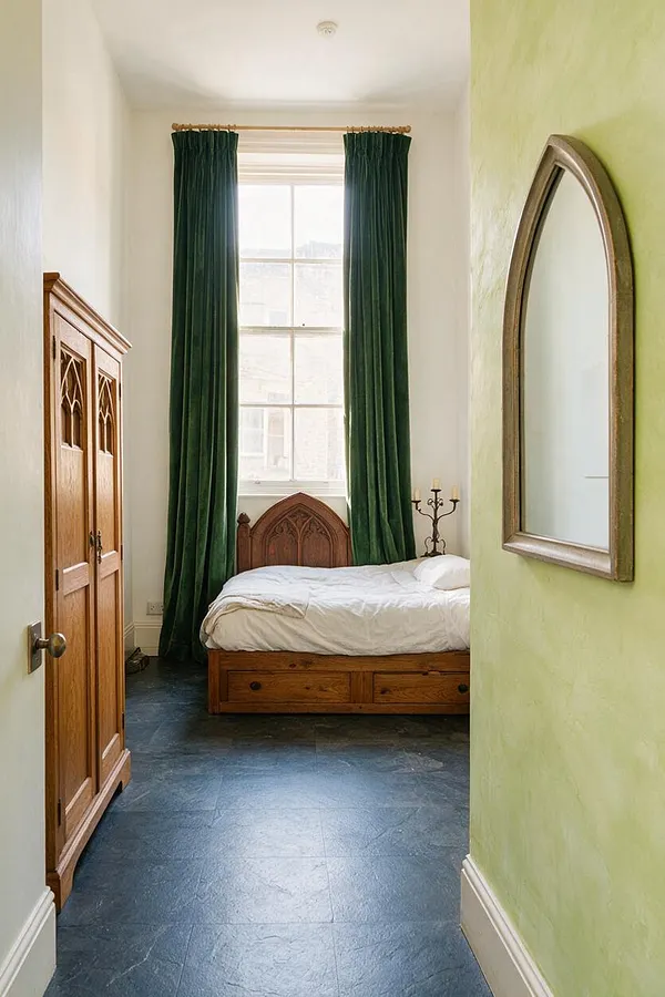A narrow studio apartment bedroom with a wooden bed and carved Gothic headboard centered beneath a tall window dressed in dark green velvet curtains. A matching Gothic-style wardrobe stands on the left, while an arched mirror hangs on a pale green wall to the right, above a dark slate floor.