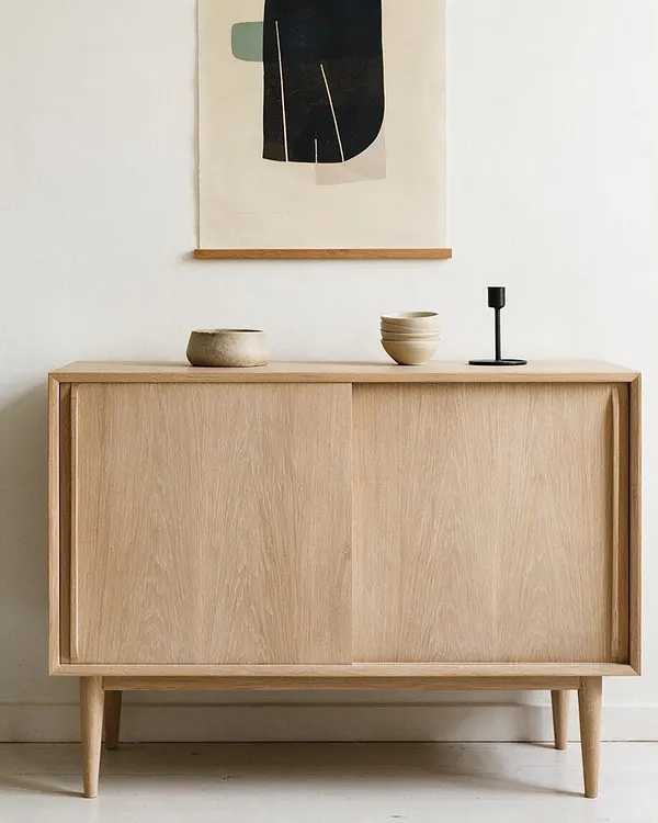 A light oak sideboard with slim tapered legs and sliding doors sits against a white wall. A simple abstract artwork hangs above it, and the top is styled with small ceramic vessels and a black candleholder.