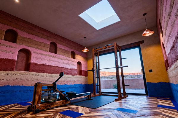 A Southwestern home gym features rammed-earth style walls in horizontal bands of adobe pink, terracotta, sand, and cobalt blue, dotted with arched wall niches. Natural light pours from a ceiling skylight and a large sliding glass door overlooking desert cliffs. A wood-framed power rack on a black rubber mat and a walnut water rowing machine sit on a herringbone wood floor with blue inlays, while hammered copper pendants add warm illumination.