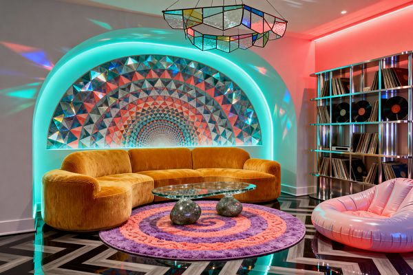 A psychedelic living room centers on a plush mustard velvet curved sectional set before an arched alcove lined with triangular iridescent mosaic tiles, haloed by neon teal backlighting and reflected by a multicolor stained-glass pendant. A round purple-and-coral spiral shag rug anchors a glass coffee table with two polished stone bases, while a pink inflatable lounge and a stainless-steel record shelf loaded with vinyls sit on a glossy black-and-gray chevron floor under pink and teal ambient lighting.
