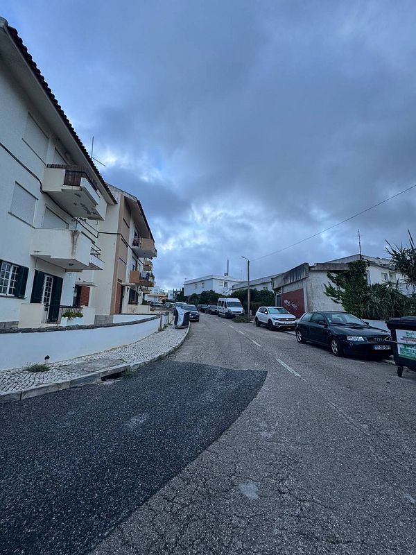 A narrow street in a beach town with parked cars and cloudy skies.