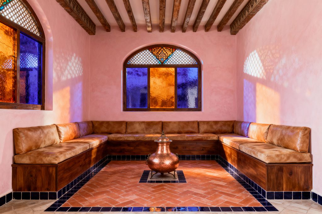 A Moroccan-style living room with blush pink plaster walls and exposed wooden ceiling beams features a U-shaped built-in wooden banquette topped with camel-colored leather cushions. The floor is terracotta laid in a herringbone pattern with a black tile border, and a hammered copper urn sits at the center. Arched windows with cobalt blue and amber stained glass and lattice details cast colorful light and patterned shadows across the room.