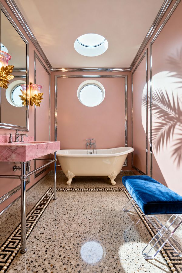 A Hollywood Regency bathroom with blush-pink walls framed in polished chrome, an ivory clawfoot tub centered under a round porthole window, and a pink marble console sink on chrome legs. Terrazzo floors with a black-and-cream Greek key and pearl-bead border lead to a royal-blue velvet bench on clear acrylic X-legs. A circular skylight and gold leaf sconces with pink glass shades illuminate the space and cast palm shadows.