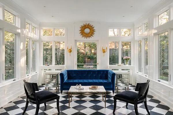 A bright sunroom with white paneled walls and many tall windows on three sides, centered around a deep blue velvet sofa. Black accent chairs, mirrored side tables, a marble coffee table, and a black-and-white diamond checkered floor create a glamorous Hollywood Regency look, with a gold sunburst mirror above the doorway.