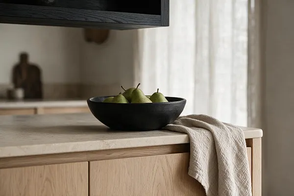 A serene Japandi kitchen vignette featuring a light wood cabinet with a pale stone countertop. A matte black bowl filled with green pears sits on the counter beside a draped beige textured cloth, with soft daylight filtering through sheer curtains in the background.