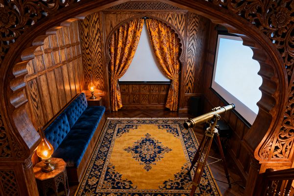 An intimate Victorian-style media room clad in carved, quarter-sawn oak paneling features a deep blue tufted velvet banquette along one wall and an arched niche with gold damask curtains revealing a projection screen. A mustard-and-navy rug sits on herringbone parquet, amber oil lamps glow on side tables, and an antique-style brass telescope stands near a second pull-down screen.