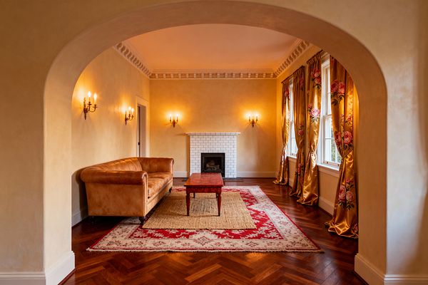 A traditional living room seen through a wide arch features warm plaster walls, dentil crown molding, and a centered fireplace clad in white subway tiles. A caramel leather sofa faces a wood coffee table on layered rugs—an ivory-and-crimson patterned rug over natural jute—while tall windows on the right are dressed in gold satin drapes with large pink roses, and brass candelabra sconces cast warm light over herringbone hardwood floors and white trim.