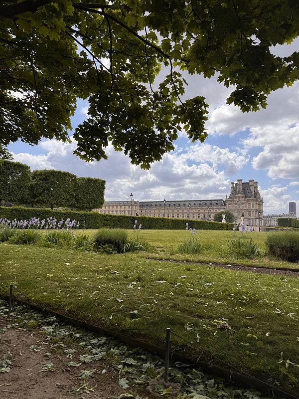 A scenic view of a park with manicured lawns, blooming flowers, and a historic building in the background under a partly cloudy sky.