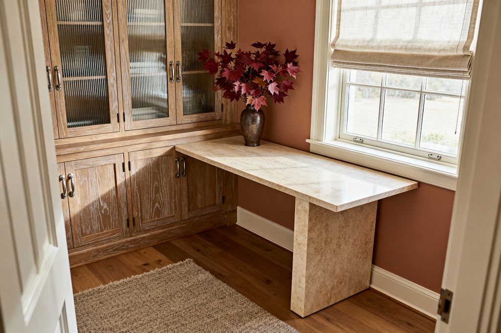 A compact home office corner shows cerused oak built-ins with reeded glass doors and metal pulls beside a polished light-beige stone desk that meets the cabinetry and sits under a window. Terracotta walls with white trim, a natural linen Roman shade, oak flooring, and a textured neutral rug create a warm, layered look; a dark bronze vase filled with deep red leaves rests on the desktop.