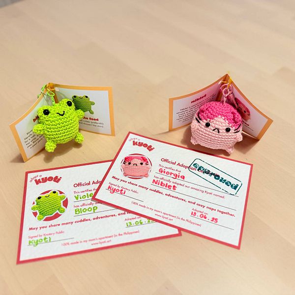 The image features two crocheted plush toys with adoption certificates on a wooden surface.