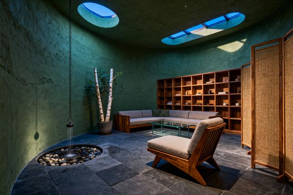 A circular living room with rough moss-green plaster walls and two oval skylights casts daylight onto dark slate tile. An L-shaped teak-framed sofa with beige cushions faces a clear glass coffee table and a matching lounge chair. A wall of open teak cubbies holds books and ceramics, a rattan folding screen stands to the right, a potted birch adds greenery, and a rain-chain fountain trickles into a recessed pebble basin.