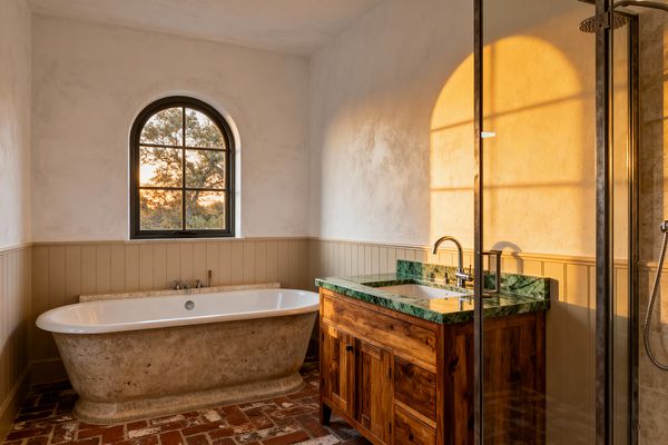 A rustic farmhouse bathroom glows with sunset light through an arched black-framed window above a freestanding stone tub set on reclaimed red brick. To the right, a solid wood vanity with a green marble countertop and chrome faucet stands against putty-beige beadboard and limewashed plaster walls, with a steel-framed glass shower in the foreground, creating a warm palette of white, terracotta, and deep green.
