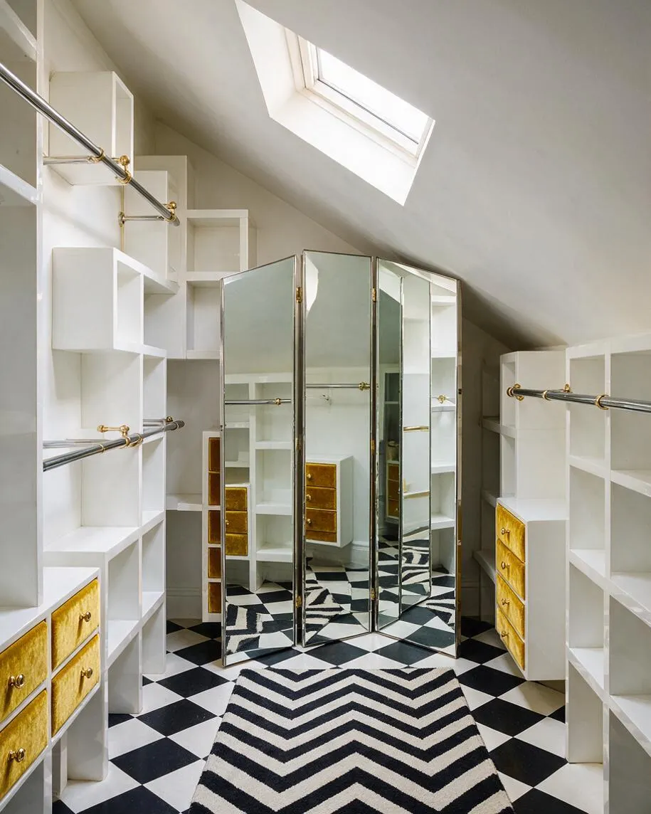 An attic closet with sloped ceilings features white built-in shelving, chrome hanging rods with brass brackets, and gold velvet-front drawers on both sides. In the center, a folding mirrored screen reflects the room, while a black-and-white diamond tile floor and chevron runner add bold Hollywood Regency flair.