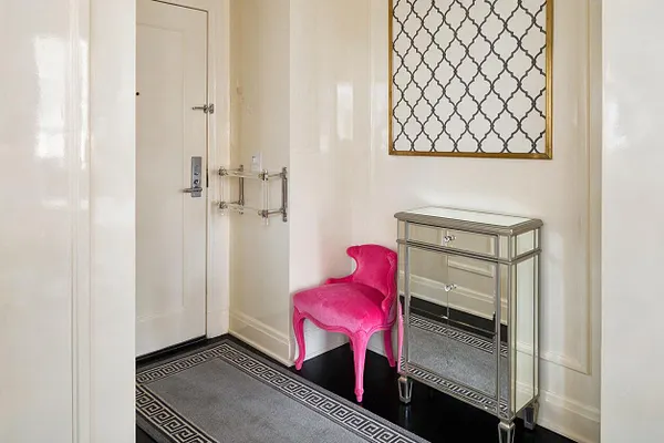 A compact Hollywood Regency entryway with glossy white walls, a mirrored console table, and a vivid hot pink velvet accent chair. A framed geometric lattice artwork hangs above, while a Greek key rug and dark floor anchor the bright, polished space.