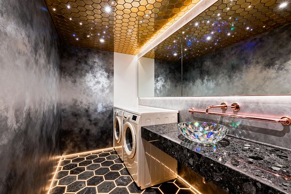 A dramatic laundry room features a gold hexagon-tiled ceiling dotted with star-like LEDs, mirrored by a long, backlit wall mirror. Smoky gray plaster walls and a glowing black hex tile floor frame a floating black terrazzo countertop with an iridescent glass vessel sink and rose-gold wall-mounted faucet; a white front-loading washer and dryer sit beneath bright task lighting, surrounded by warm ambient LED strips.