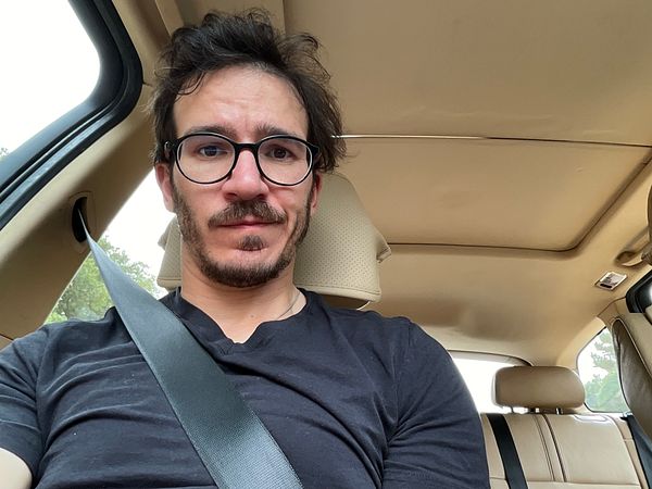 A man is seated in a car, wearing a black shirt and glasses, with a serious expression.