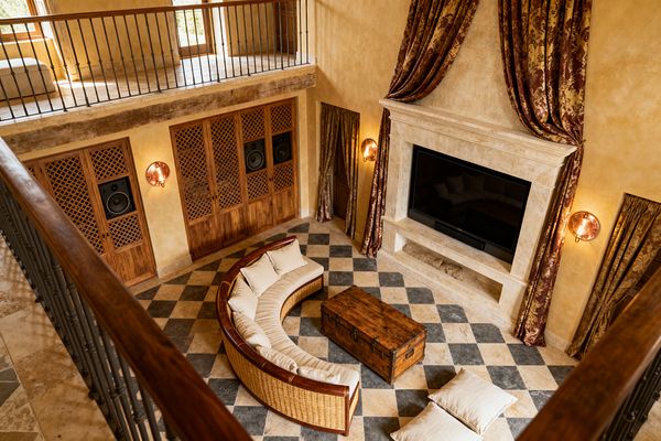 Double-height French Country media room viewed from an upper railing, featuring a carved limestone fireplace with a large flat-screen TV, burgundy-and-gold damask drapery, and warm plaster walls. A curved rattan sectional with cream linen cushions surrounds a rustic wood trunk on a tumbled limestone and charcoal slate checkerboard floor, with latticed wood cabinets and in-wall speakers, copper-toned sconces, floor pillows, and wrought-iron balustrades adding texture and warm light.