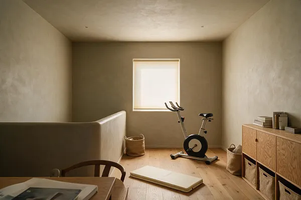 A minimalist home gym with a stationary bike centered beneath a small window, soft beige plaster walls, and pale wood flooring. Woven baskets, a low wood storage cabinet with fabric bins, and a folded exercise mat add natural textures to the calm, neutral space.