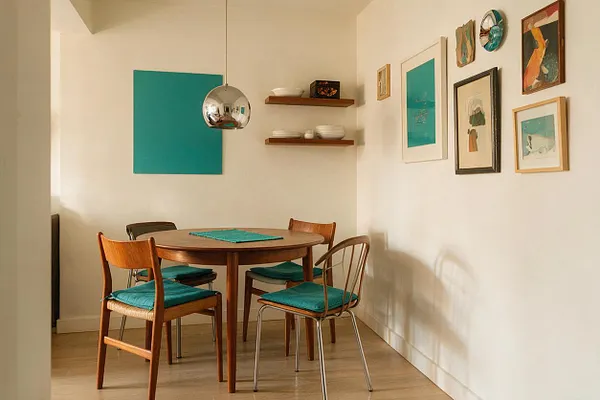 A compact eclectic dining room with a round wooden table surrounded by four mixed-style chairs, each with teal seat cushions. White walls hold a large teal abstract panel, floating wood shelves with dishes, and a gallery of colorful framed art, while a chrome globe pendant hangs overhead.