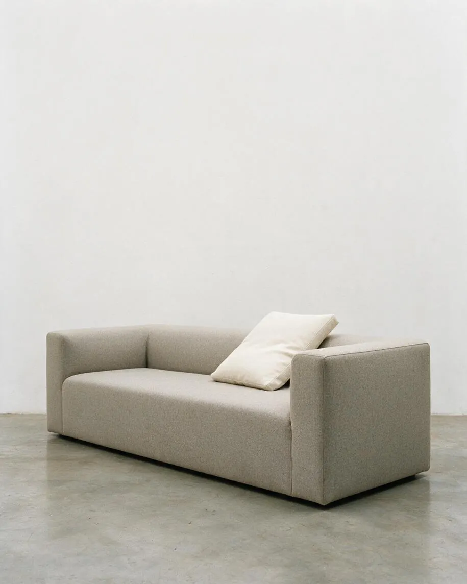 A low, boxy beige sofa sits against a plain white wall on a polished concrete floor. It has straight arms, a smooth upholstered finish, and a single off-white cushion leaning on the right side, creating a simple minimalist look.