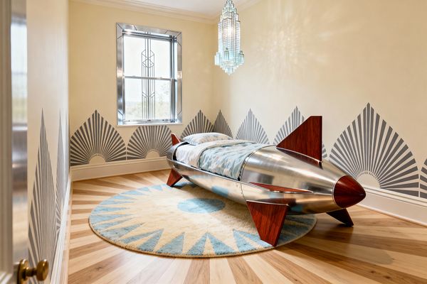 A compact kids’ bedroom with cream walls stenciled in gray Art Deco fan motifs and a light herringbone wood floor. A polished metal rocket-shaped bed with reddish wood fins rests on a round cream and pale aqua sunburst rug beneath a tiered glass chandelier. A single window with a shiny metal frame and geometric muntins brightens the space, while soft blue patterned bedding adds a playful touch.
